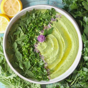 Bowl of green goddess hummus garnished with herbs, oil and chive flowers.