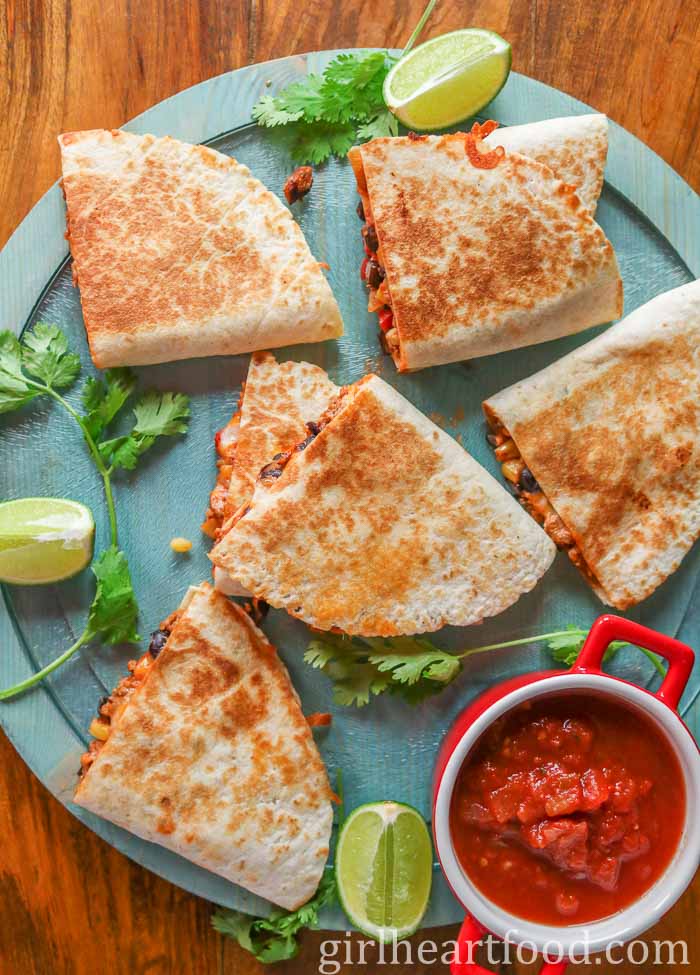 Quesadillas on a blue board next to cilantro, lime wedges and a dish of salsa.