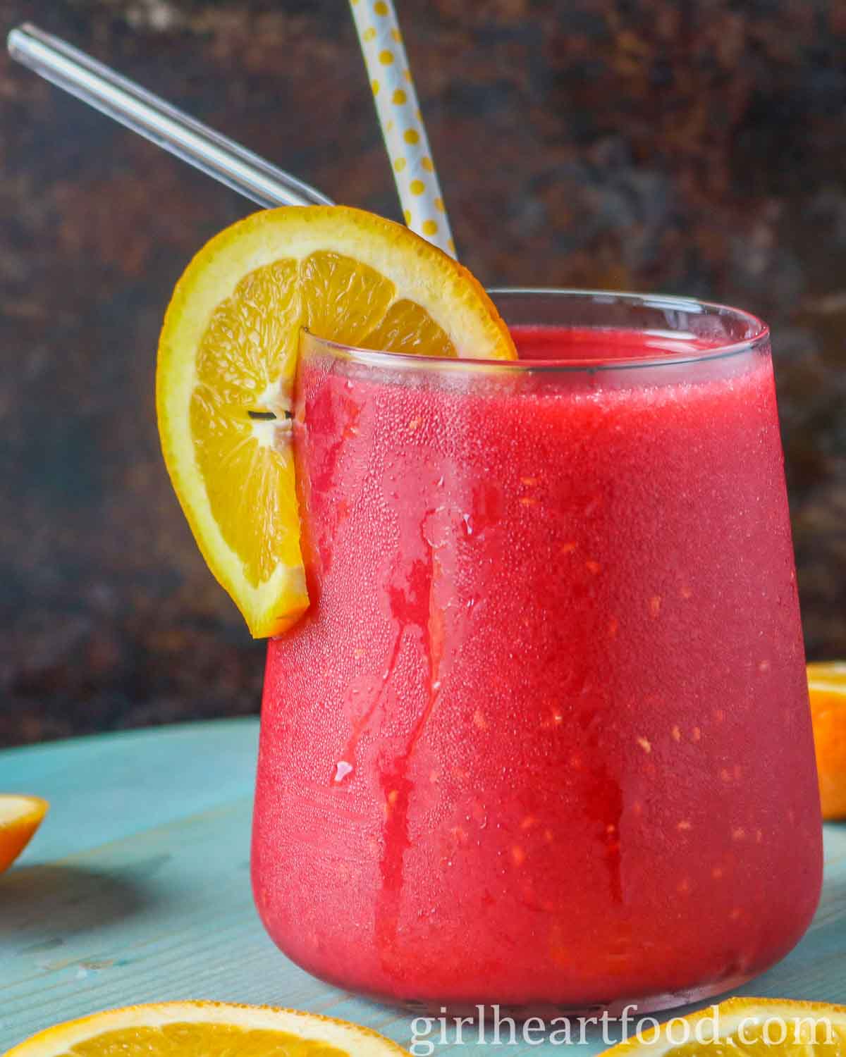 Glass of raspberry vodka slushy garnished with an orange slice.