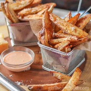 Two baskets of crispy potato wedges next to a dish of dipping sauce.