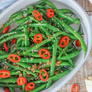 Garlicky green beans in a dish topped with sliced chili pepper and sesame seeds.