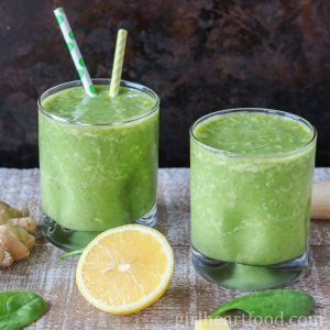 Two glasses of green smoothie alongside spinach, cut lemon and ginger.