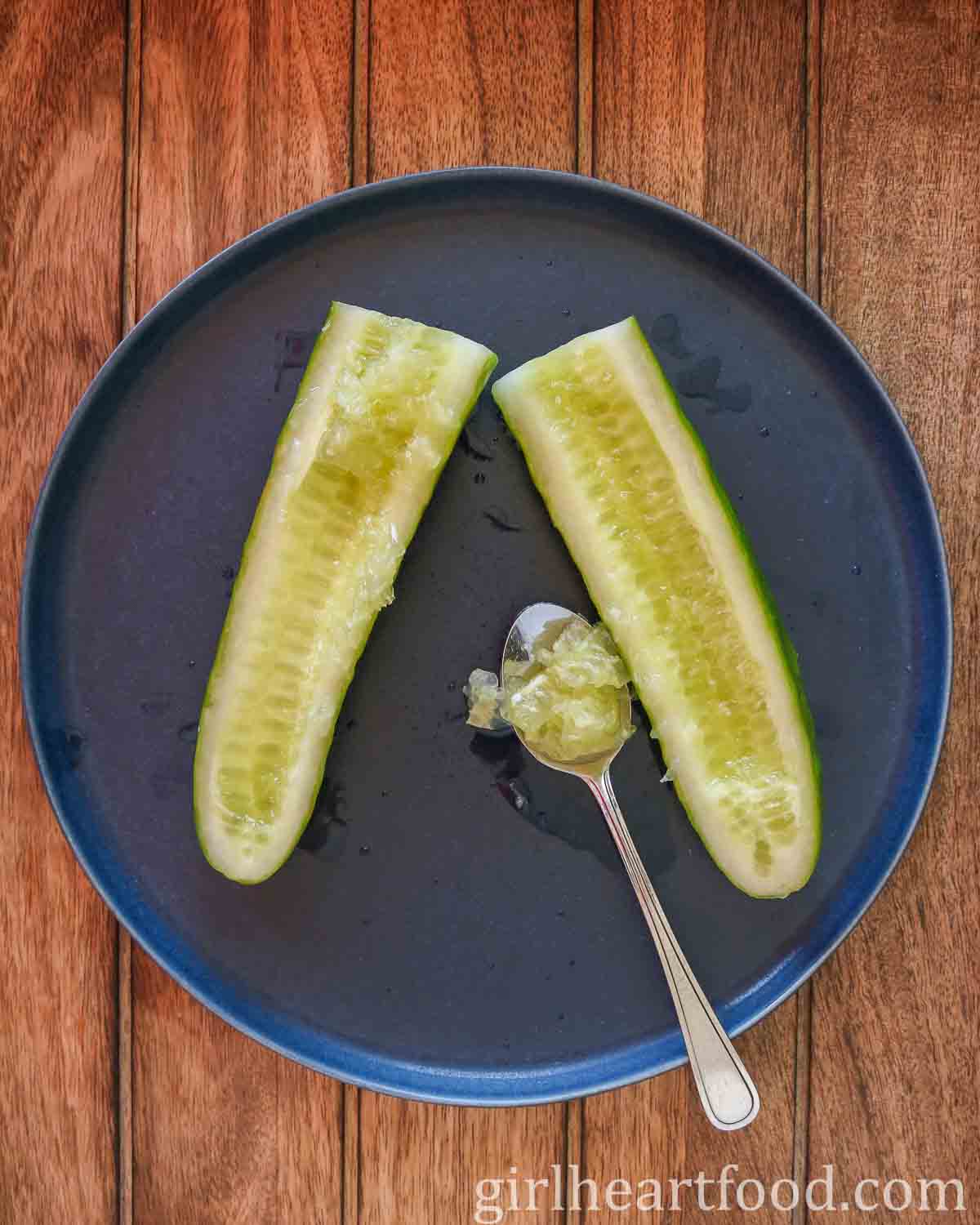 Two halves of a cucumber on a blue plate with the interior seeds scooped out with a spoon.