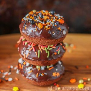 Stack of three pumpkin chocolate donuts with ganache & sprinkles alongside sprinkles.