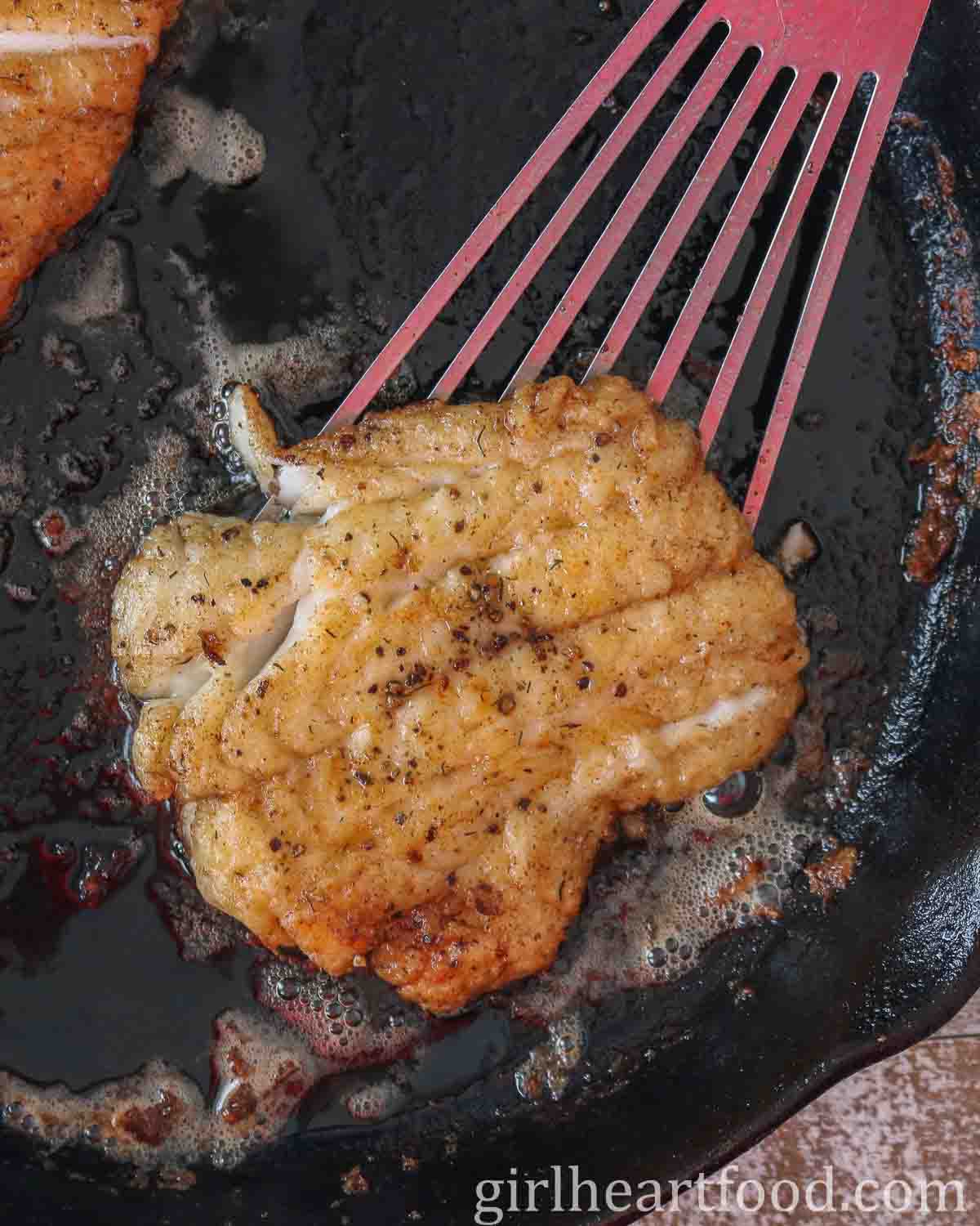 A spatula picking up a piece of cooked cod from a cast-iron skillet.