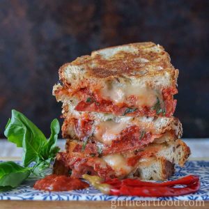 Stack of three pizza grilled cheese sandwich halves next to chili peppers, basil & pizza sauce.