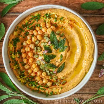 Bowl of pumpkin hummus garnished with toppings and surrounded by fresh sage.