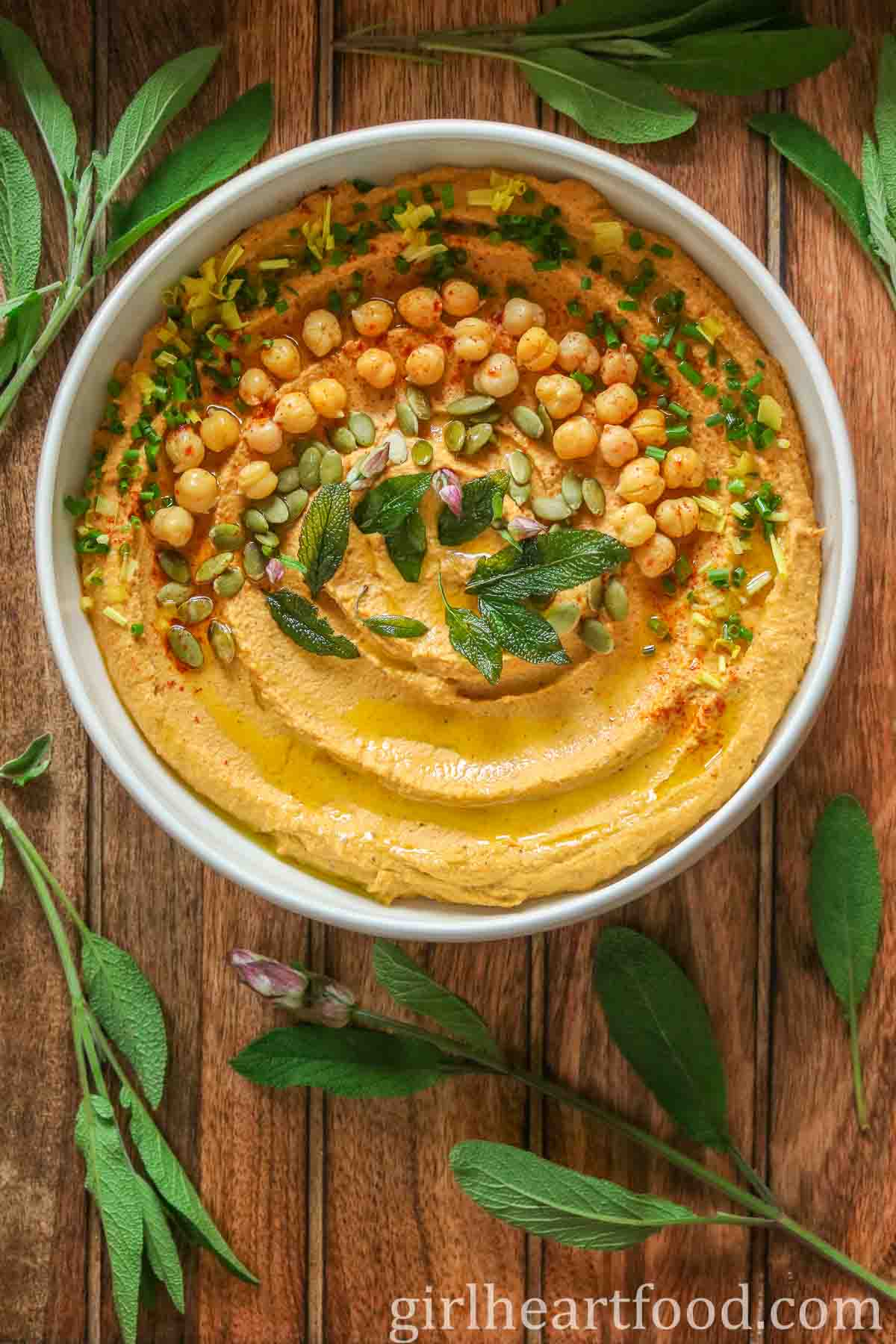 Bowl of pumpkin hummus garnished with toppings and surrounded by fresh sage.
