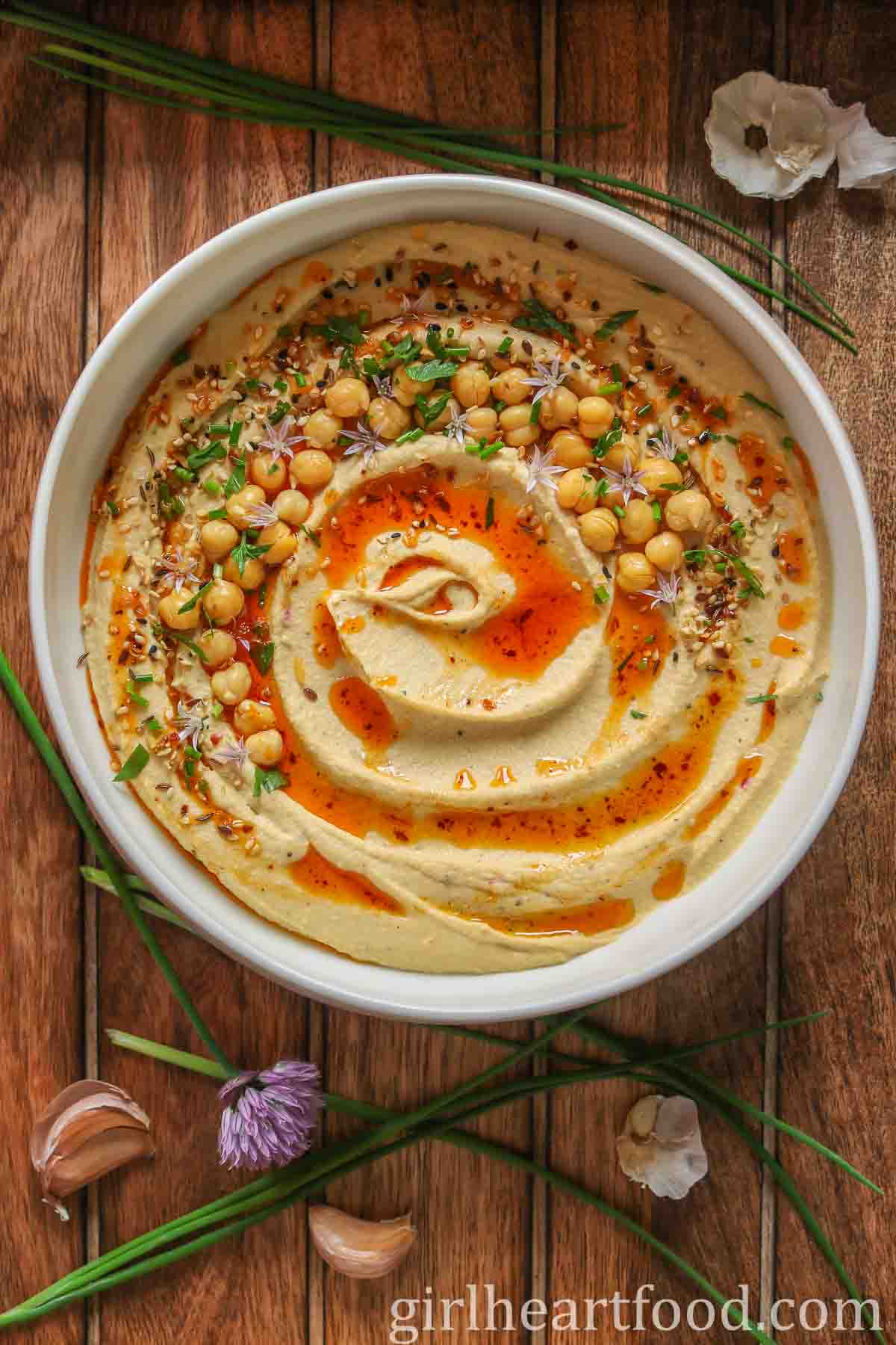 Bowl of roasted garlic hummus garnished with toppings, next to chives and garlic cloves.