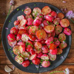 Roasted radishes garnished with chives and chive flowers on a black plate.