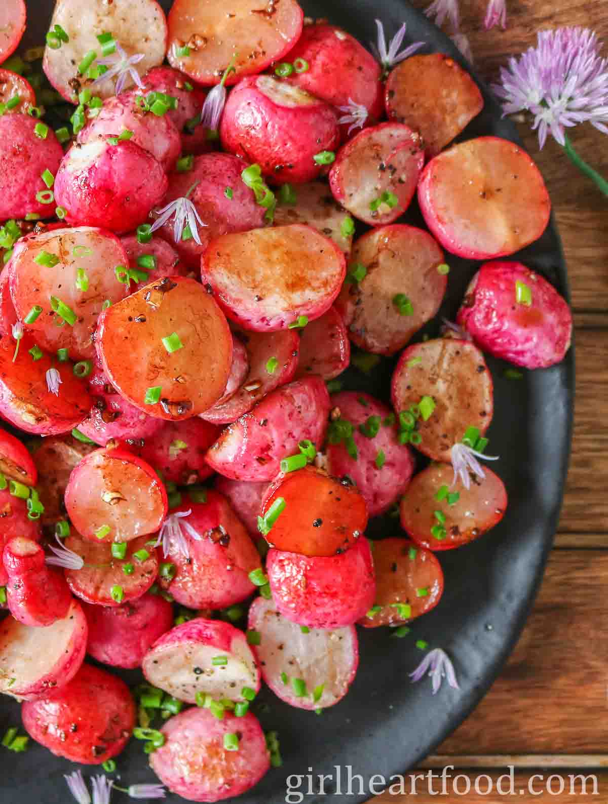 Close-up of roasted radishes garnished with chives and chive flowers on a black plate.