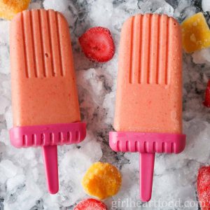 Two strawberry mango kefir ice pops and chunks of frozen fruit on a tray of ice.