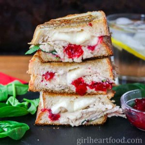 Stack of three cheesy turkey cranberry sandwich halves next to cranberry sauce & spinach.