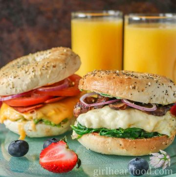 Two bagel breakfast sandwiches next to berries and glasses of orange juice.