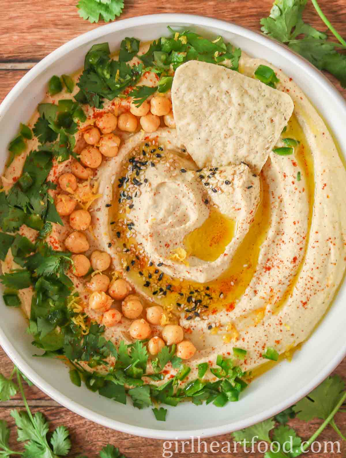 Bowl of hummus garnished with toppings and a tortilla chip dunked into it.