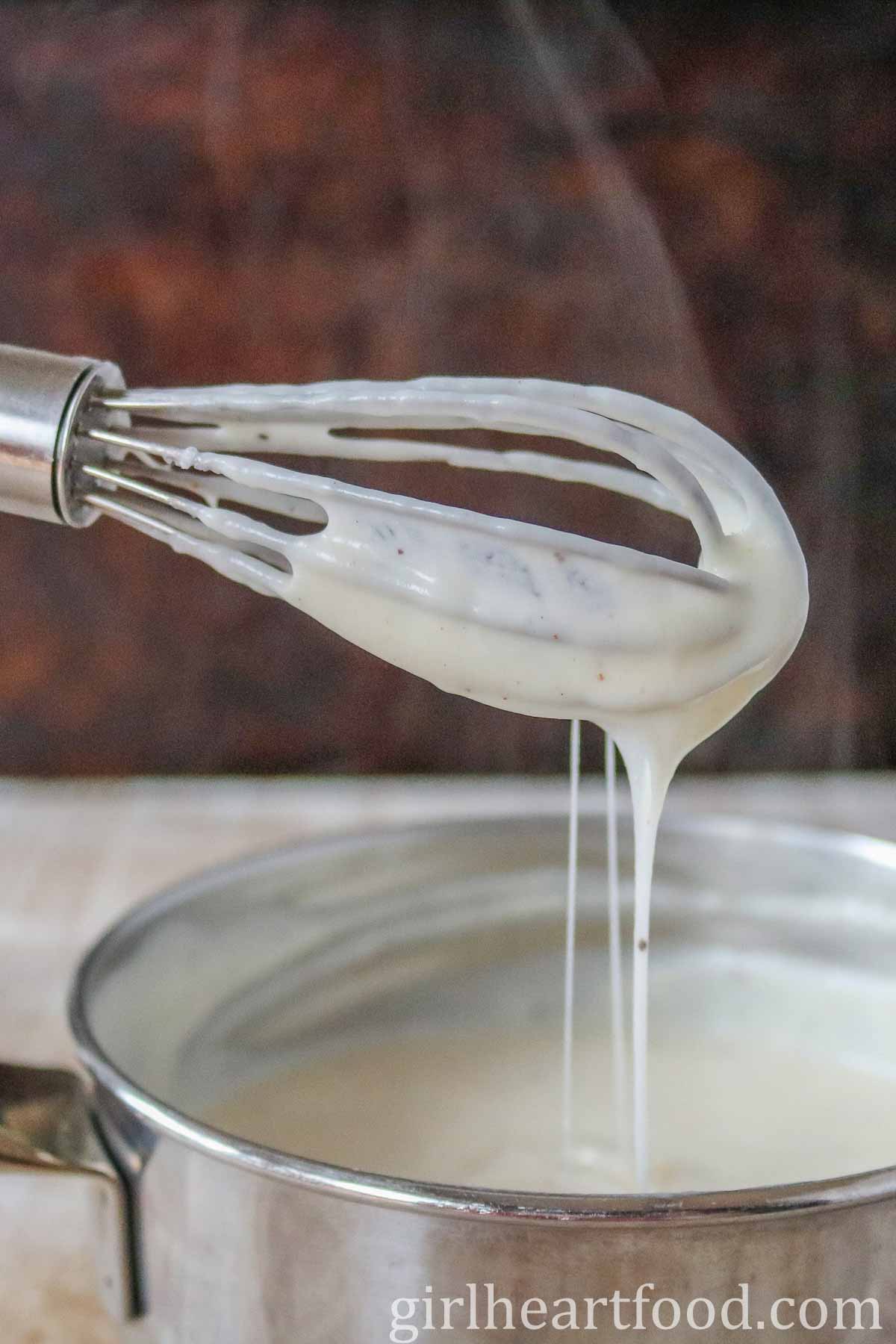 A whisk dripping with white sauce into a saucepan.