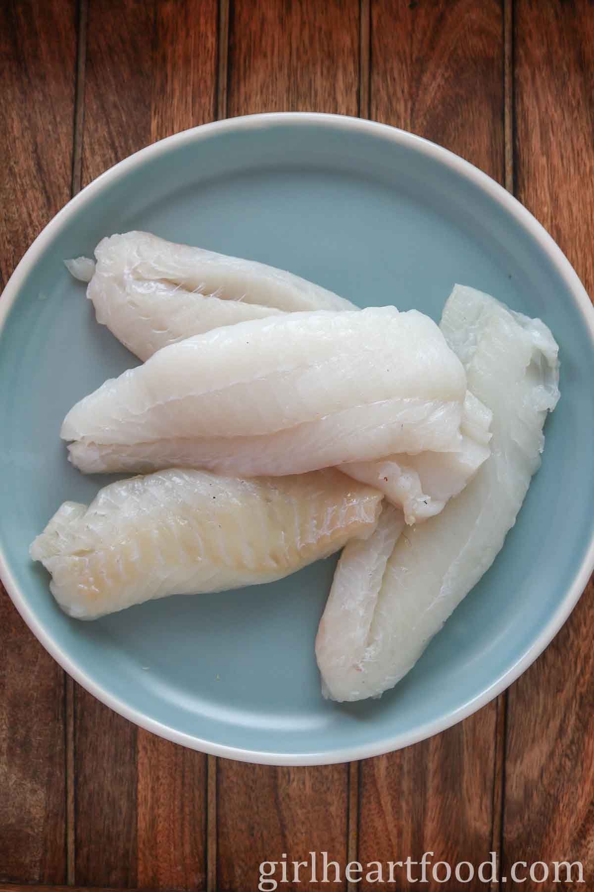 Four uncooked cod fillets on a light blue plate.