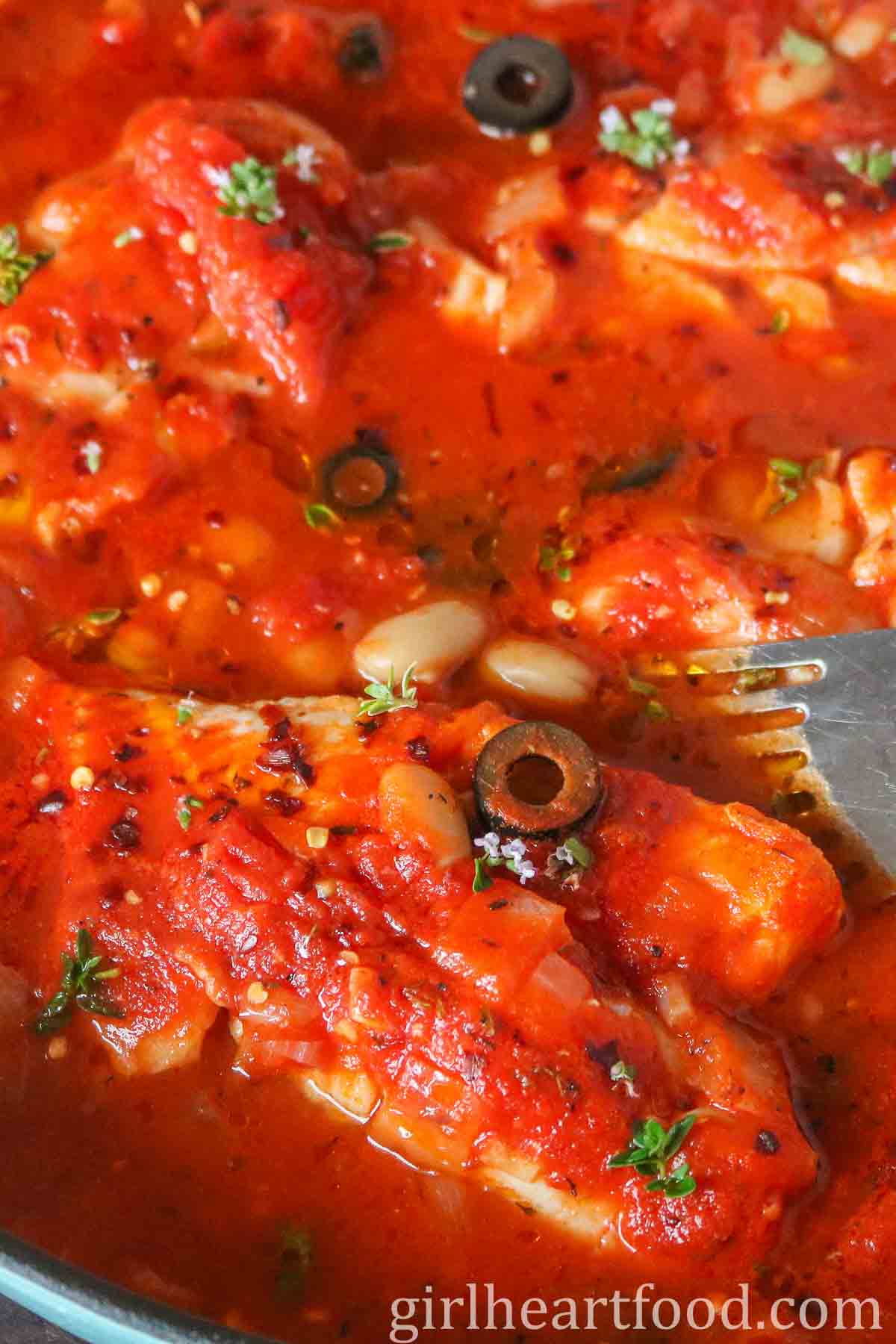 Spatula picking up a fillet of cod in tomato sauce from a pan.