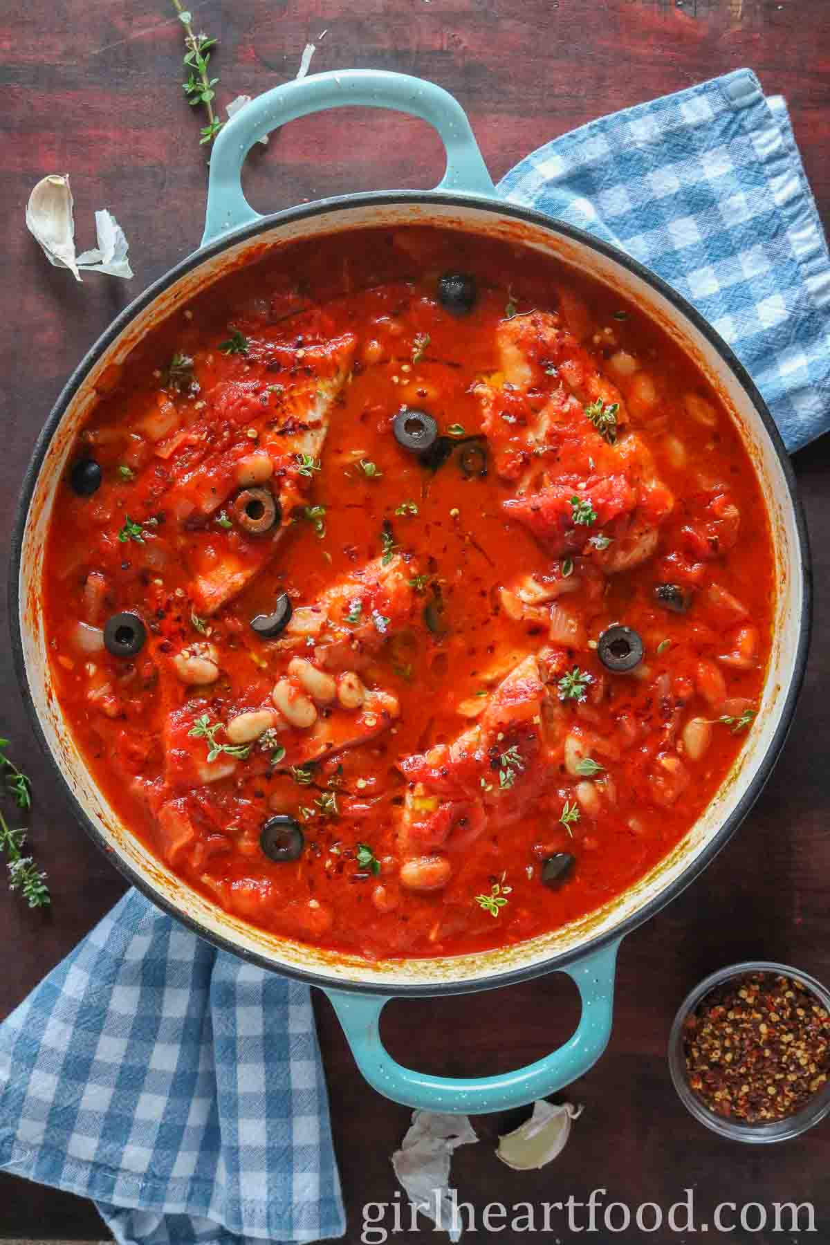 Pan with cod in tomato sauce next to a dish of crushed red pepper flakes and garlic cloves.