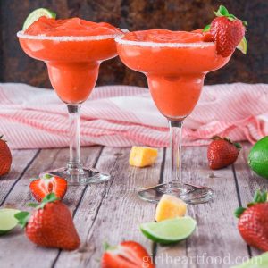 Two glasses of frozen margaritas next to lime, strawberries and cubes of mango.