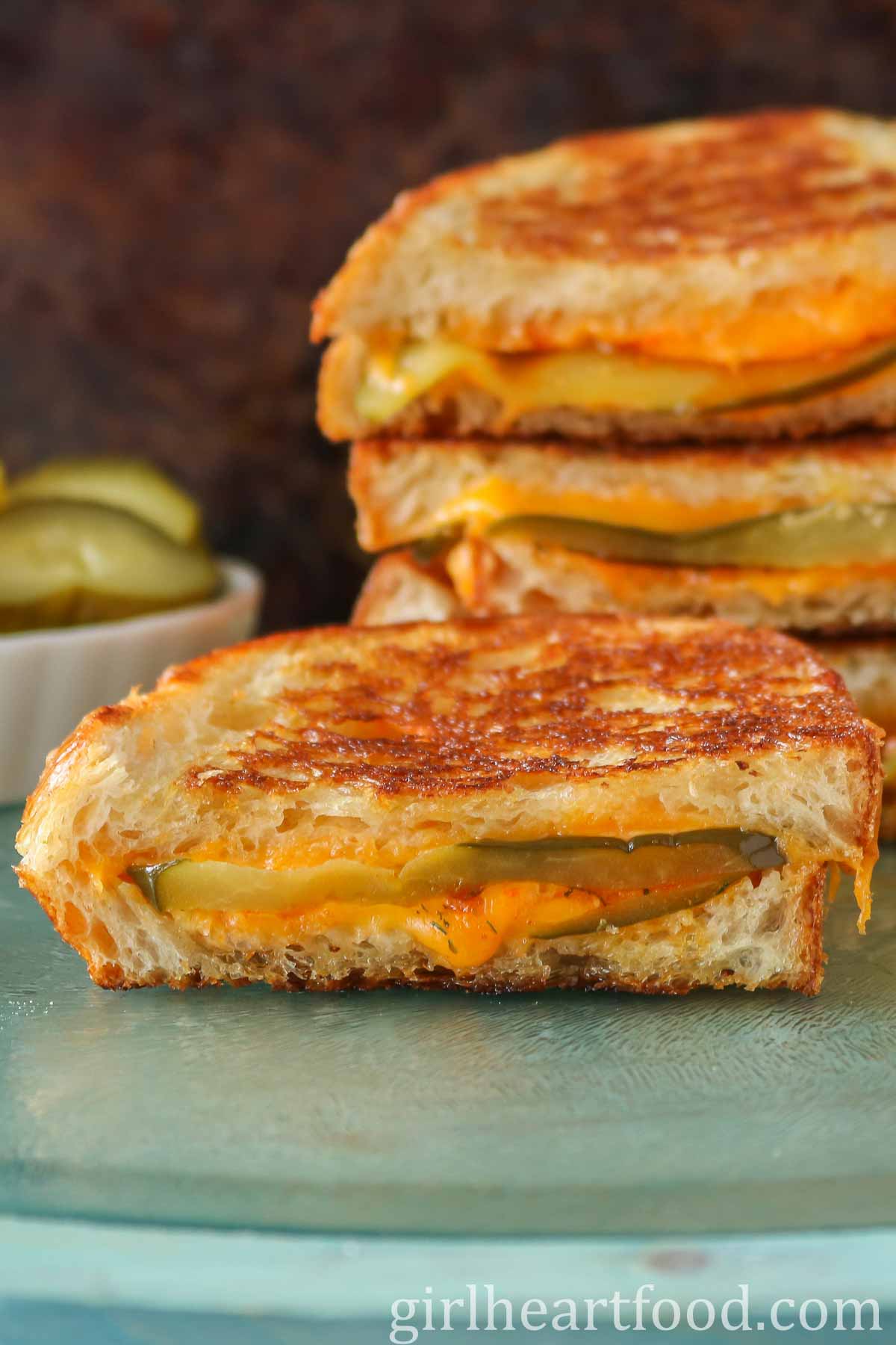 Half of a pickle grilled cheese sandwich in front of a stack of sandwiches & dish of pickles.