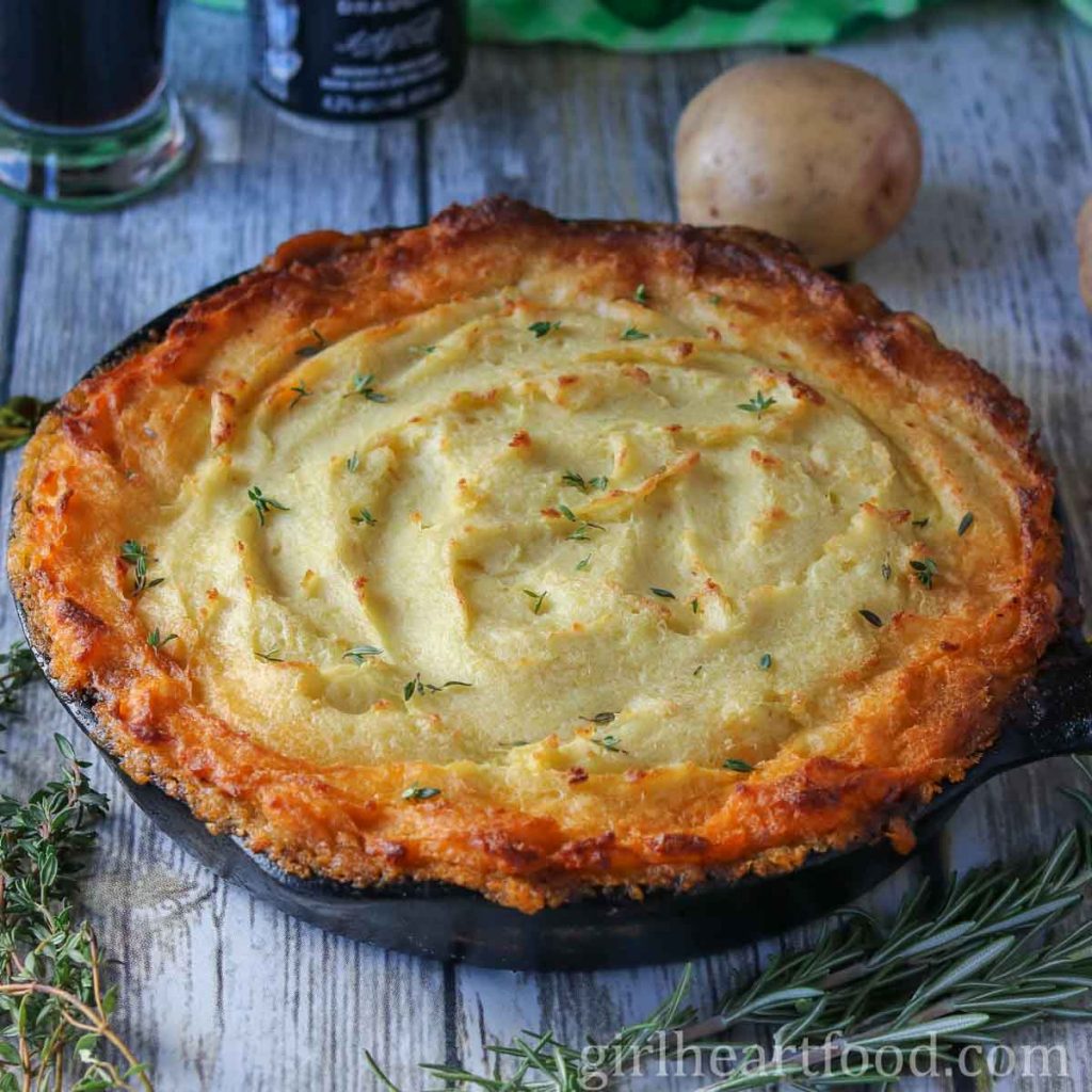 Skillet of traditional shepherd's pie next to rosemary, thyme and a potato.
