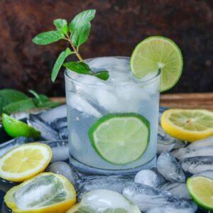 Glass of lime vodka soda garnished with mint, surrounded by ice and slices of lemon and lime.