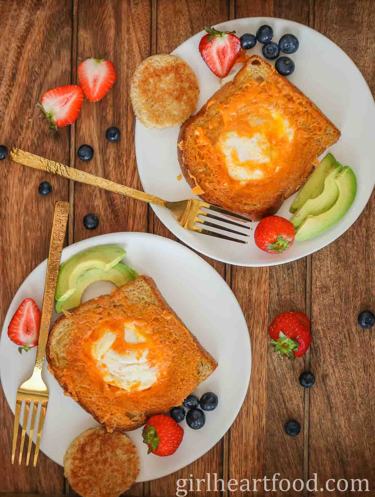 Two white plates, each with egg-in-the-hole toast, berries, sliced avocado and a gold fork.