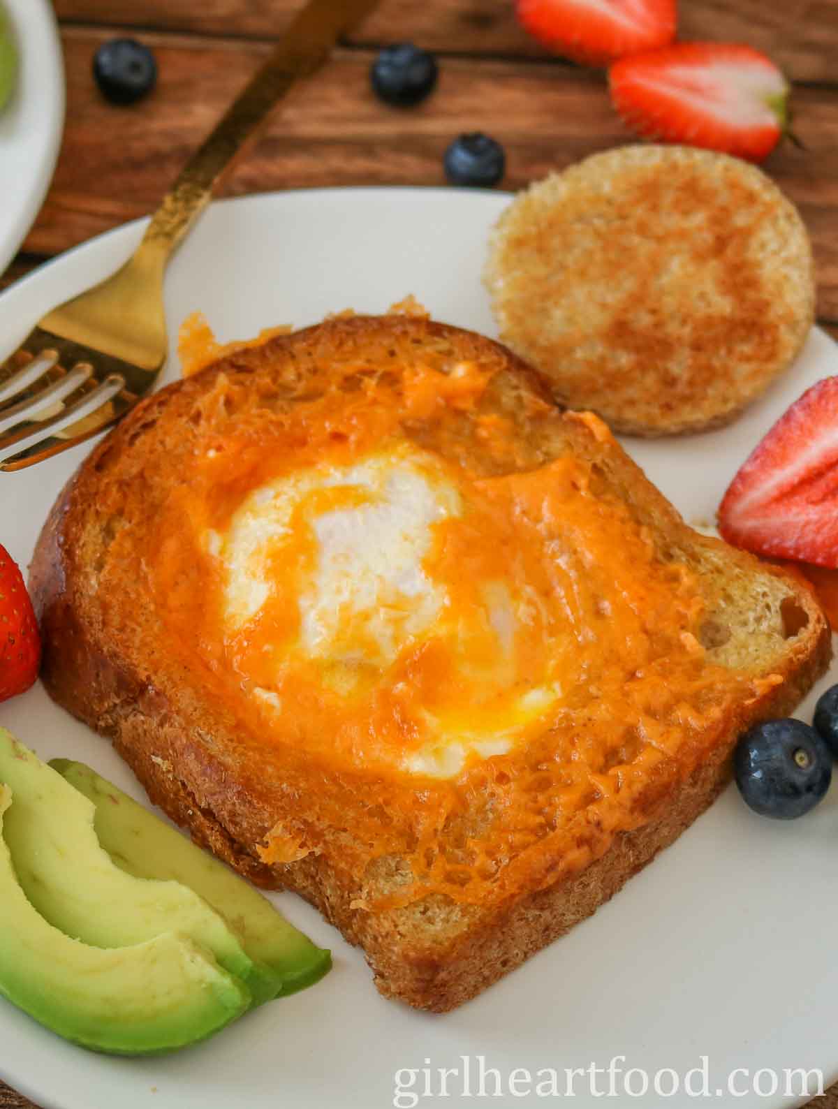 Egg-in-a-hole toast on a white plate, next to berries, sliced avocado, and a gold fork.