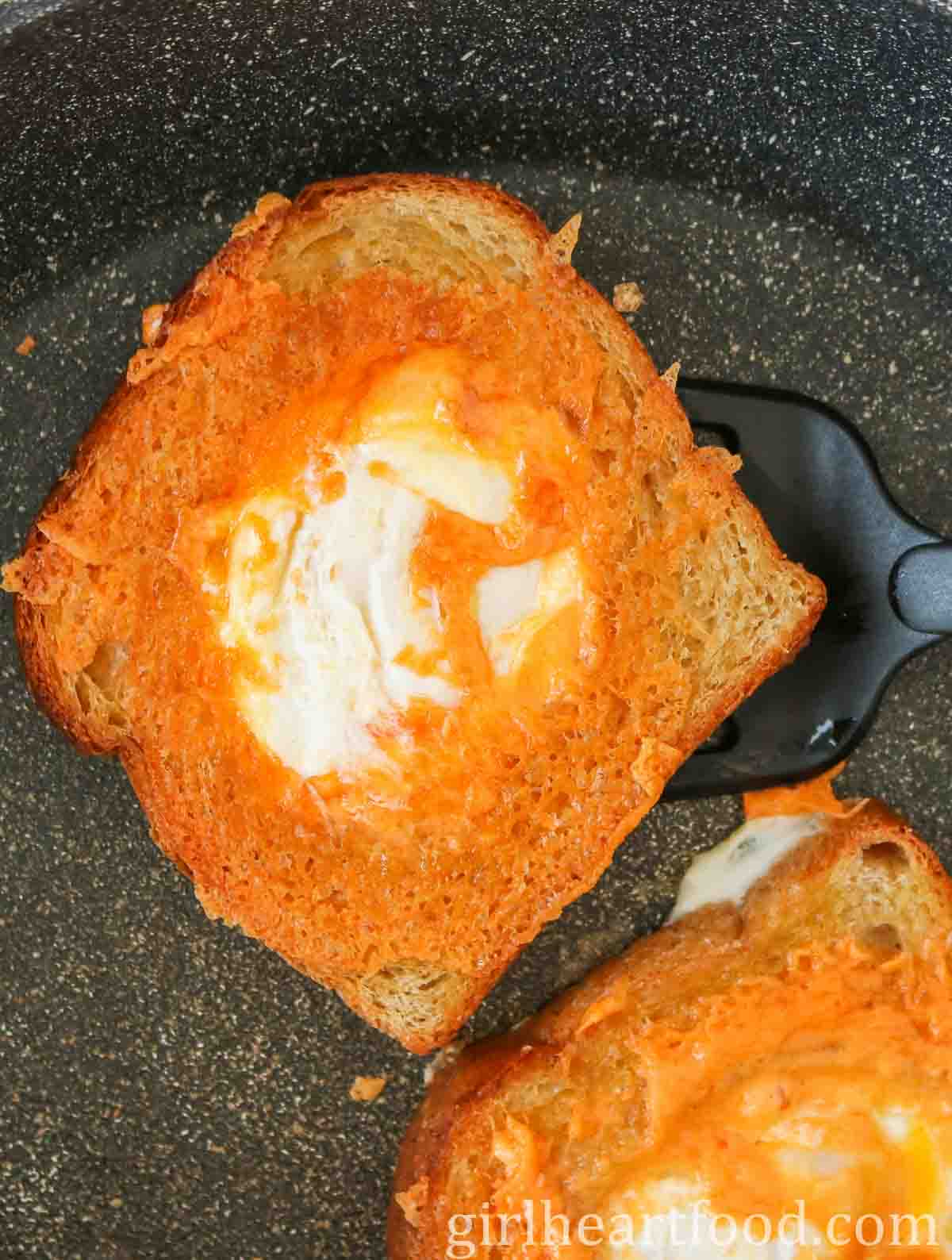 Spatula picking up cheesy egg toast from a frying pan.