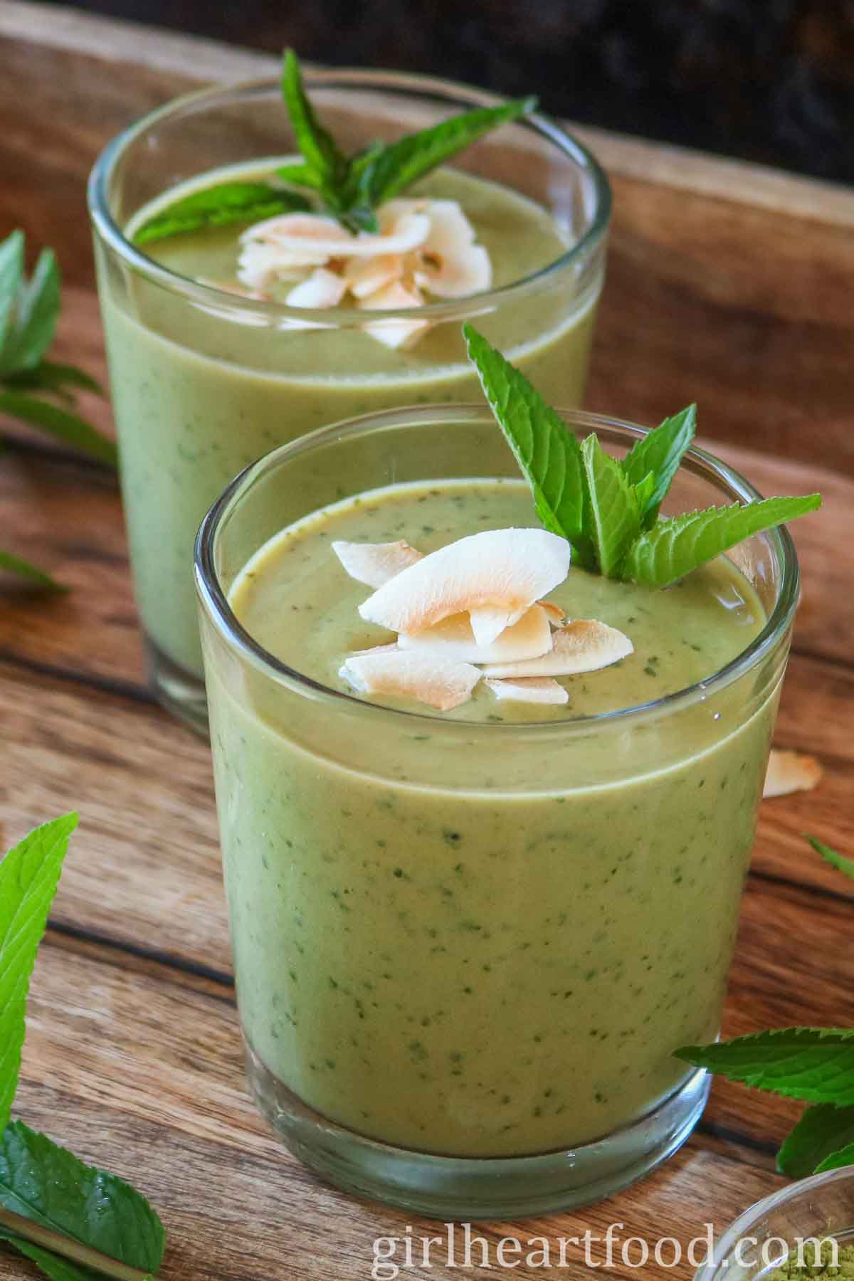 Two glasses of matcha smoothie, one in front of the other, each garnished with mint and coconut.