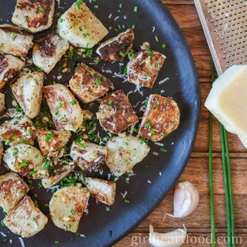 Chunks of roasted kohlrabi on a black plate garnished with grated cheese and chives.