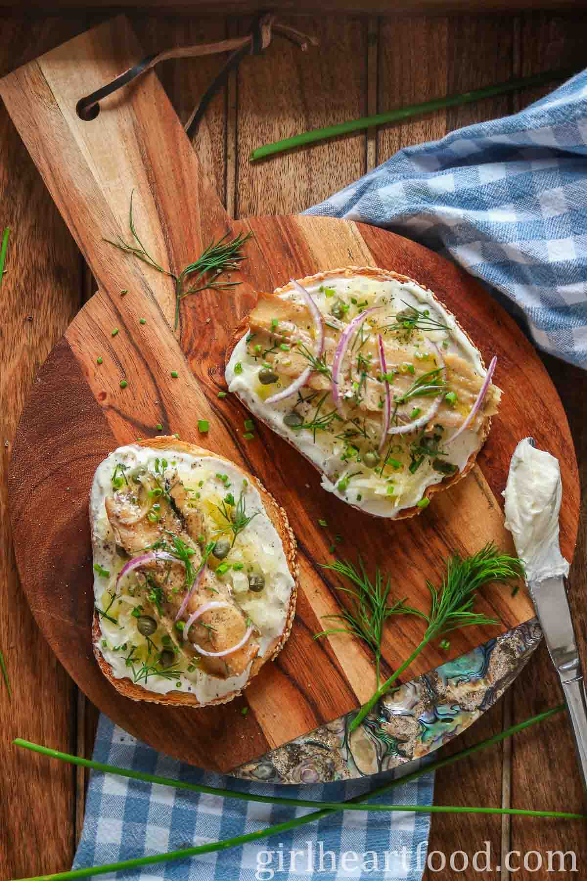 Two smoked herring and cream cheese toasts next to herbs and a knife with cream cheese on it.