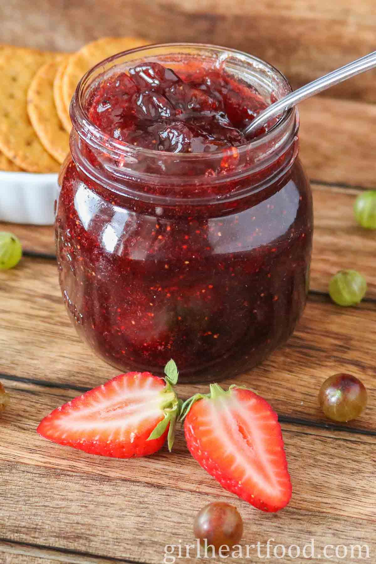 Jar of strawberry gooseberry jam with a spoon in it, next to some berries.