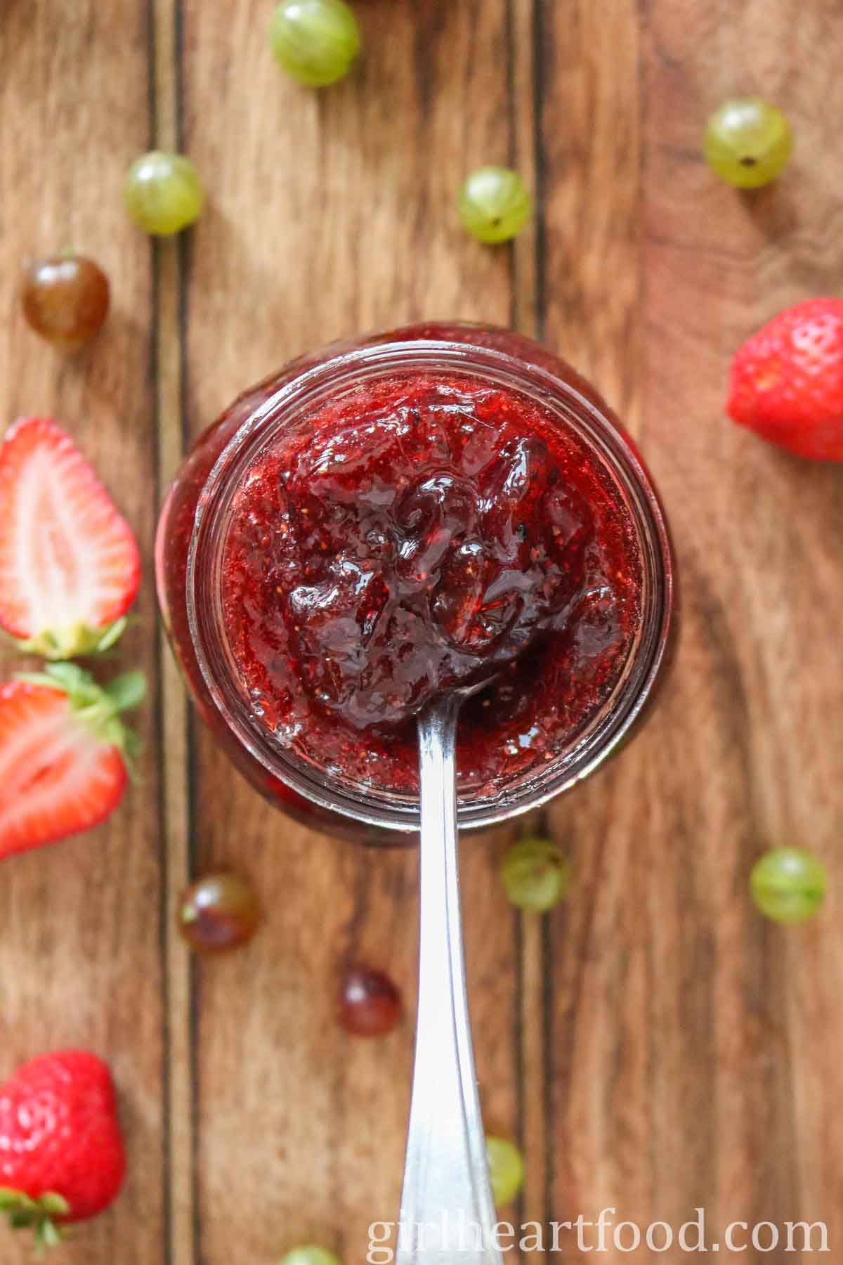 Overhead shot of a jar of gooseberry strawberry jam with a spoon dunked into the jam.