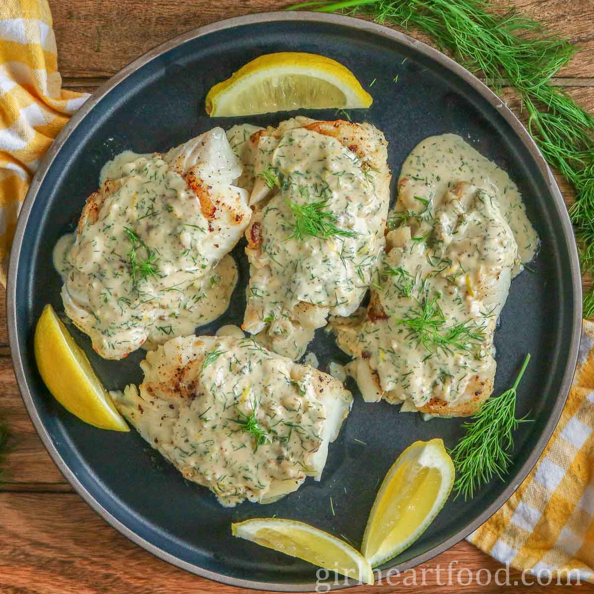 Four cod fillets with creamy dill sauce and lemon wedges on a dark blue plate.