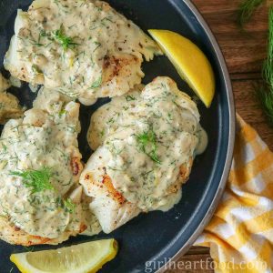 Three cod fillets with creamy dill sauce and lemon wedges on a dark blue plate.