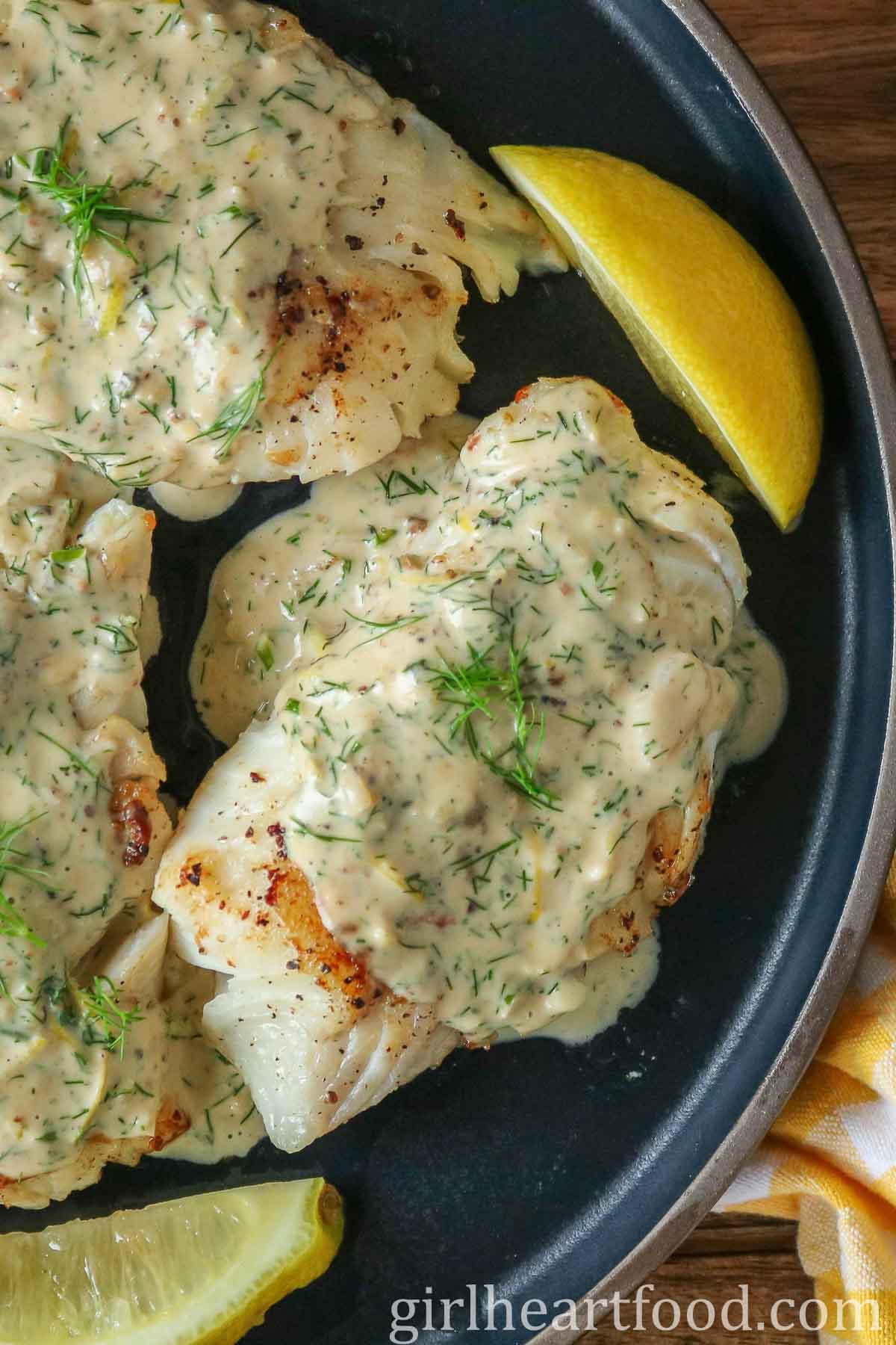 Close-up of cod fillets with a creamy dill sauce and lemon wedges on a dark blue plate.