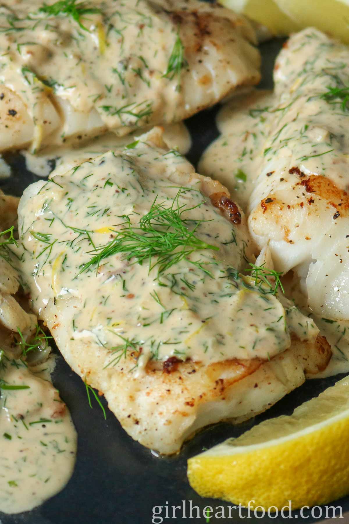 Close-up of a cod fillet covered in a creamy dill sauce next to a lemon wedge.