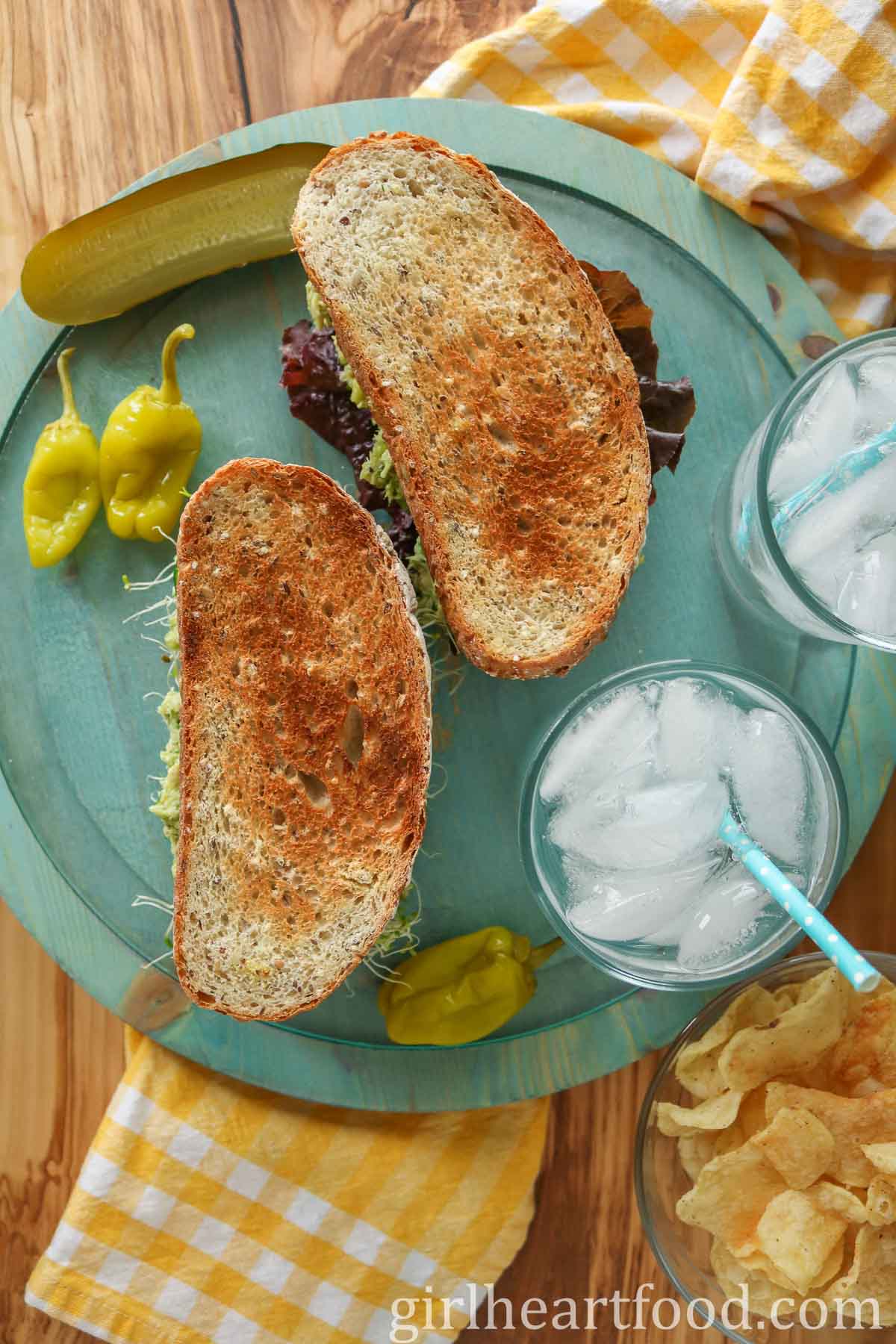 Two toasted sandwiches next to glasses of water, hot peppers, pickle and chips.