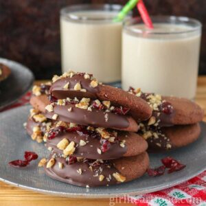 Stack of chocolate shortbread cookies with more cookies and two glasses of milk behind it.