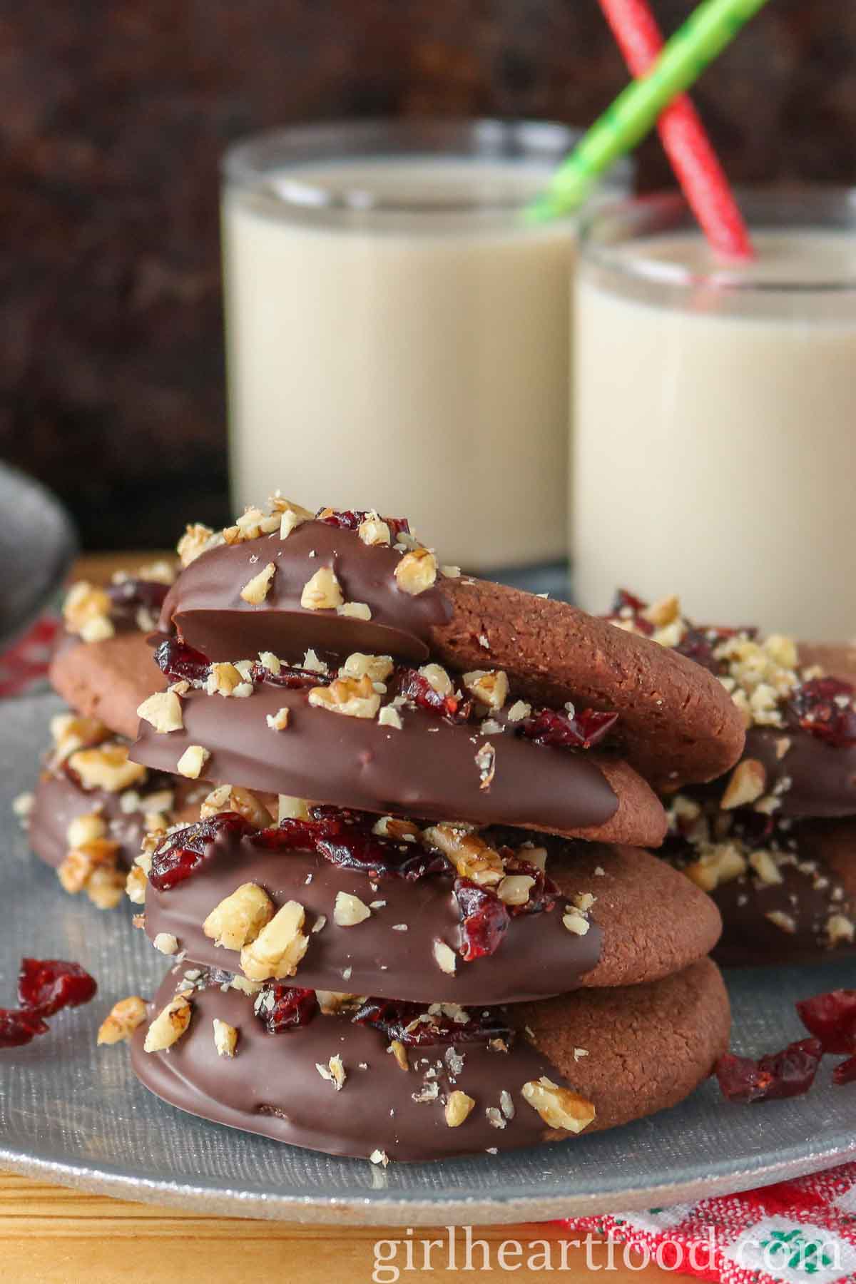 Stack of chocolate shortbread cookies with more cookies and two glasses of milk behind it.