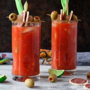 Two glasses of Caesar cocktail, each garnished with a snack and celery stick.