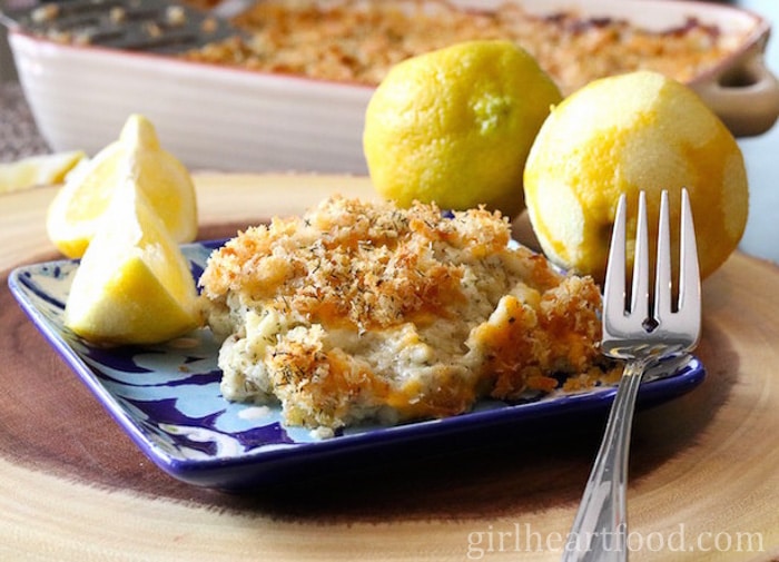 Portion of cod au gratin and a lemon wedge on a plate with a fork resting on the plate.