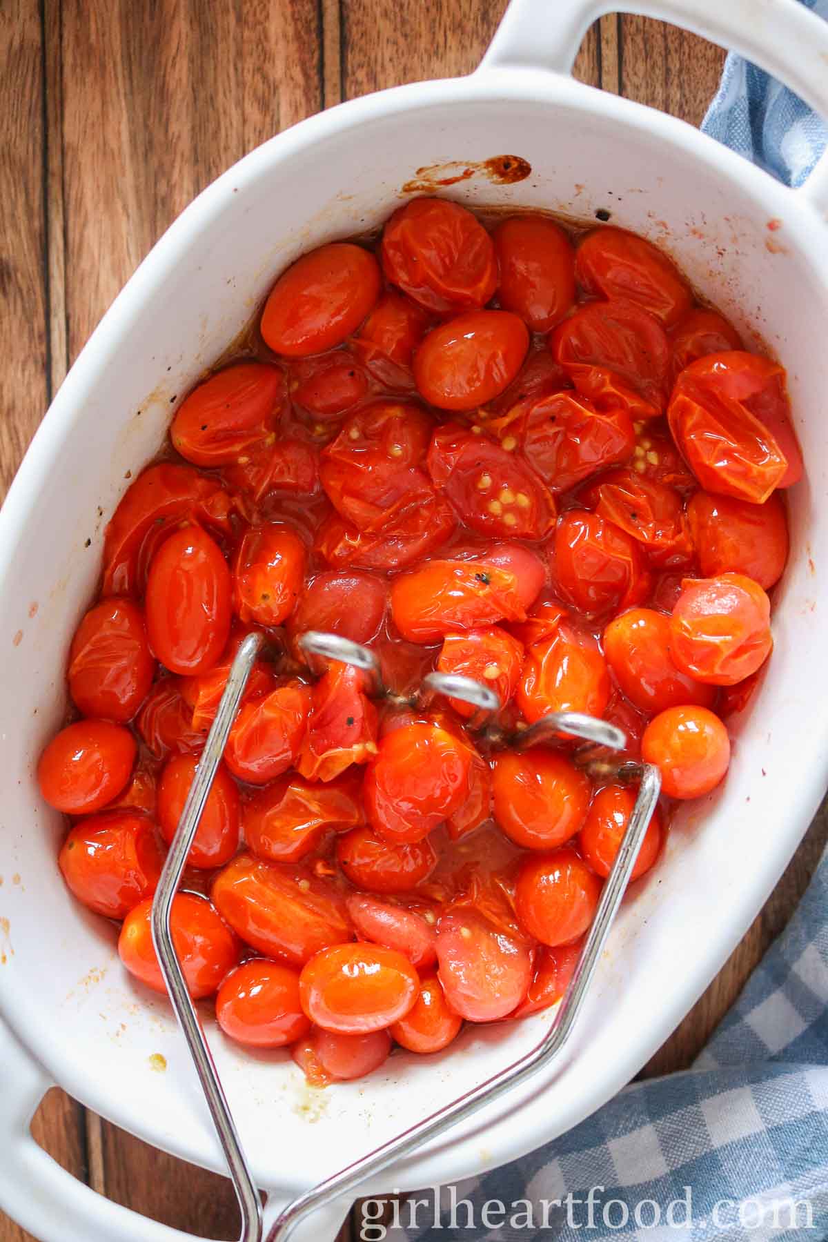 Roasted grape tomatoes in a baking dish being smashed with a potato masher.