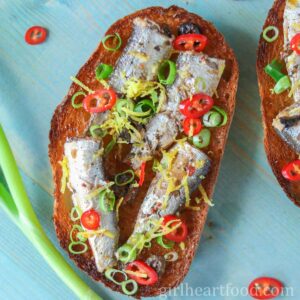 Sardines on toast with chili pepper, lemon zest and green onion next to a stalk of green onion.