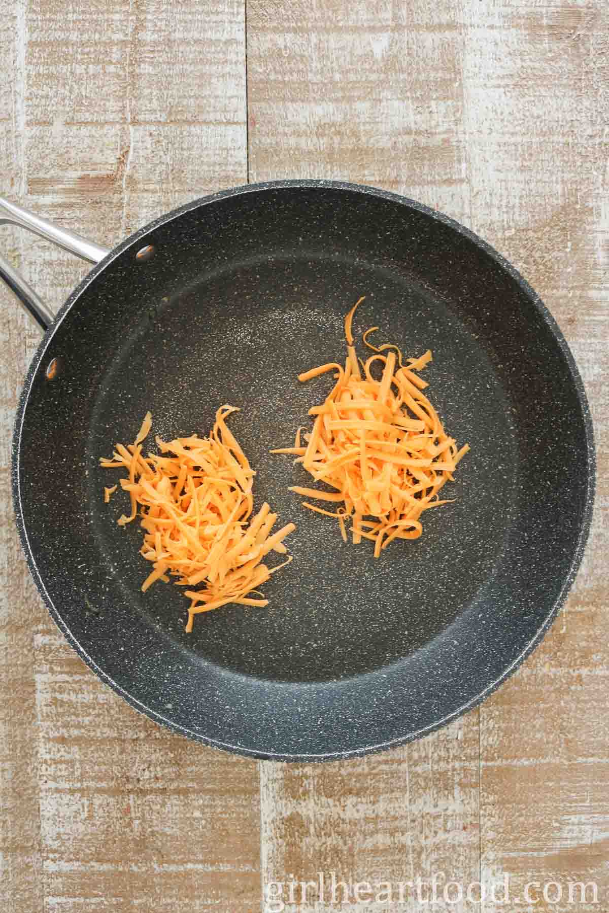 Two mounds of shredded cheddar cheese in a frying pan.