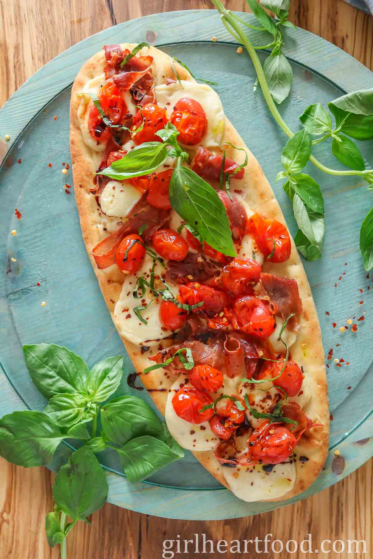 Tomato and prosciutto flatbread pizza with fresh basil on either side of it.