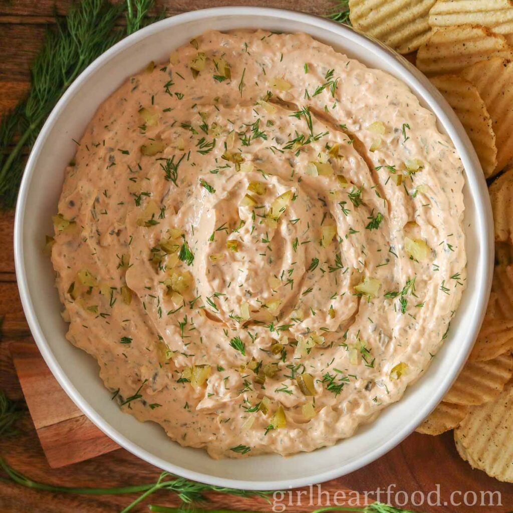 Bowl of dill pickle dip alongside fresh dill and potato chips.