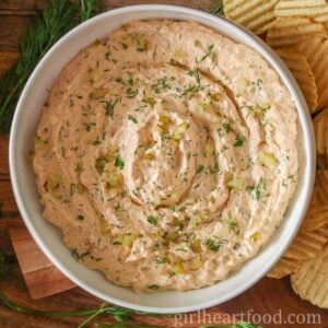Bowl of dill pickle dip alongside fresh dill and potato chips.